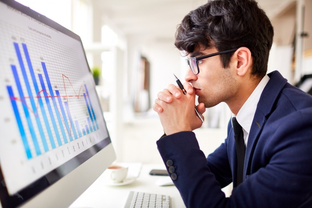 A man deep in thought, staring at a computer screen while reviewing financial reports and company data. He is analyzing the company’s financial health to make an informed career decision.