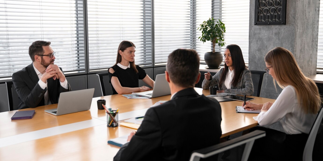 Executives sit around a table during a meeting, while a secretary writes board meeting minutes.