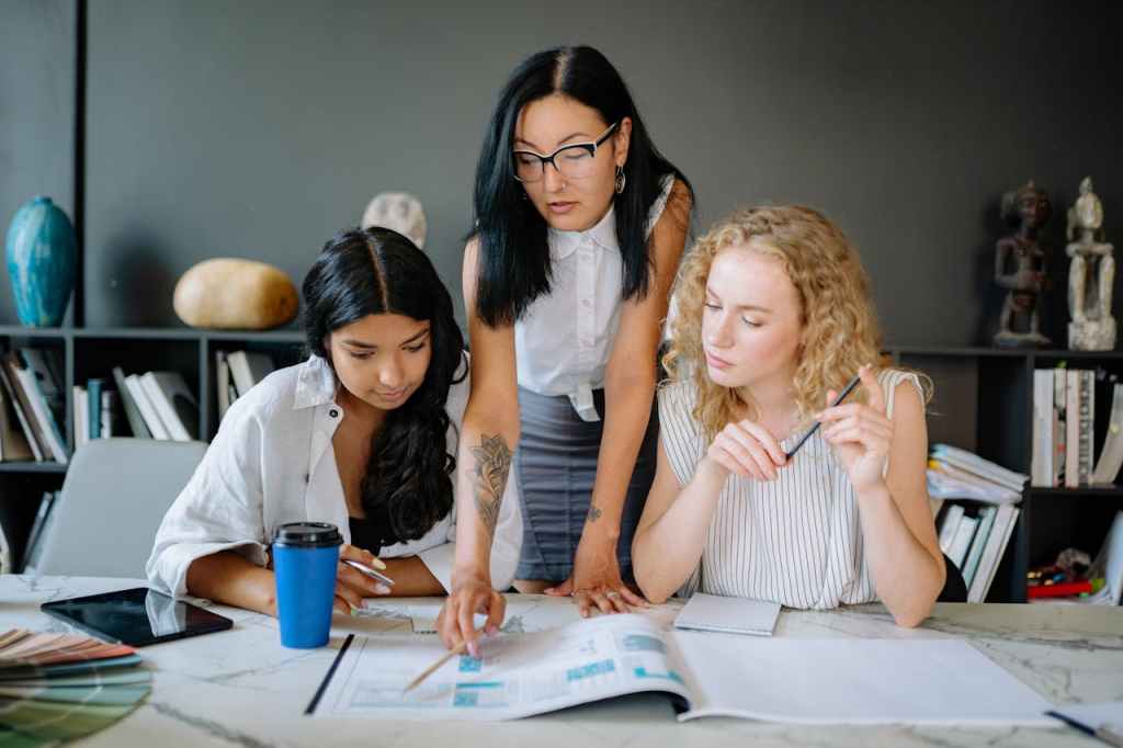 An executive assistant with a visible arm tattoo and eyeglasses is explaining a project to two employees sitting between her, demonstrating a professional and inclusive workplace environment.