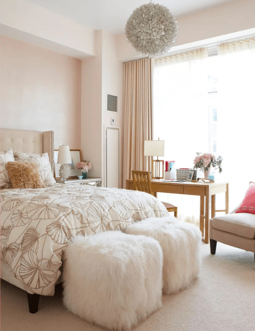 A serene white bedroom with a plush white ottoman, elegant curtains, and a simple desk area tucked into a corner.