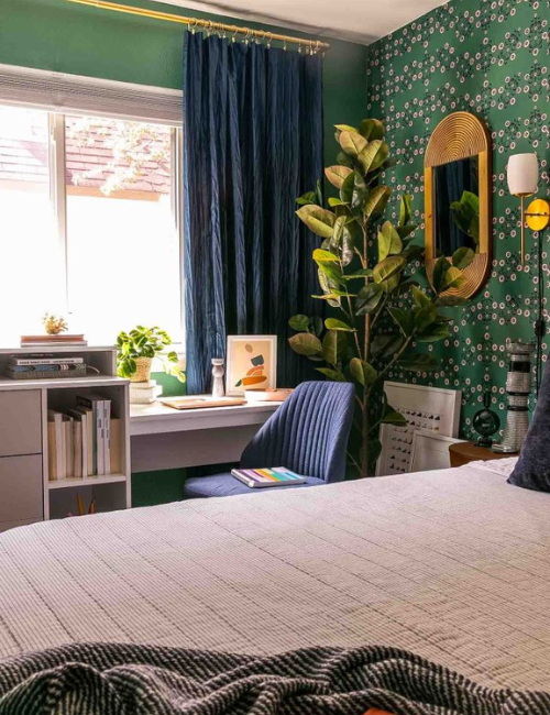 A cozy bedroom featuring dark green wallpaper with a botanical pattern, navy curtains, and a compact desk area.