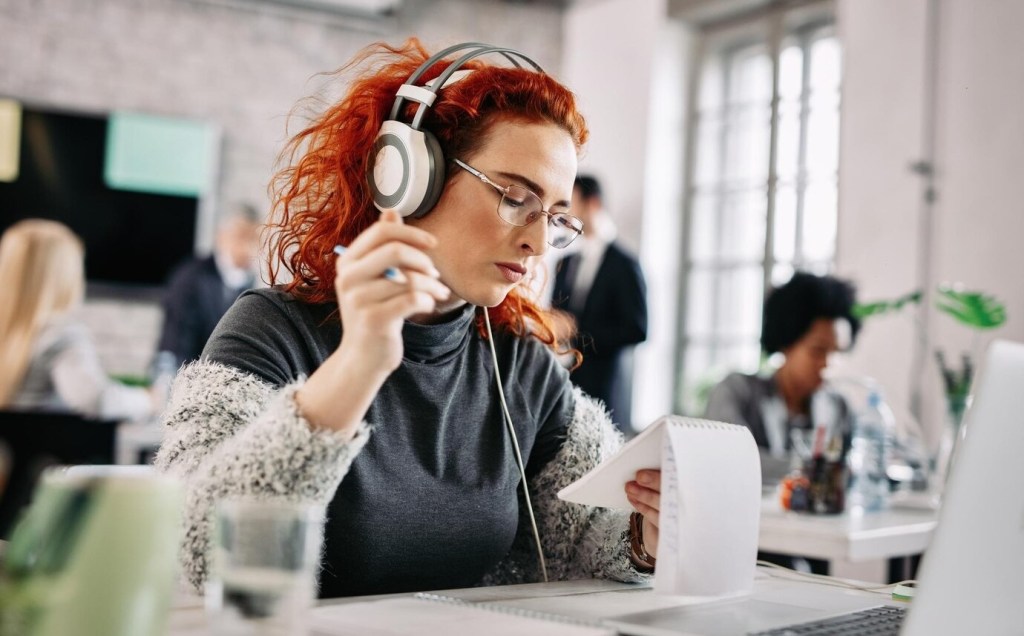 A woman wearing headphones listens to music while working in a shared office space, staying focused and respectful of her colleagues.