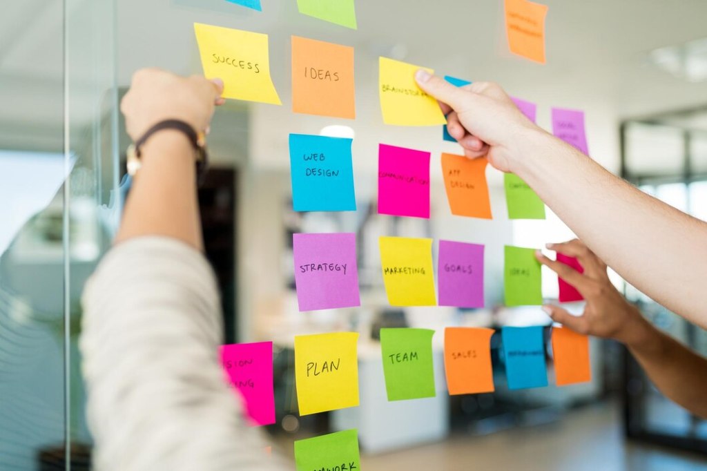 Hands arranging colorful sticky notes on a glass wall during a brainstorming session, with notes labeled with concepts like 'Success,' 'Ideas,' 'Strategy,' and 'Web Design' - demonstrating how sticky notes facilitate visual collaboration and organization of thoughts.