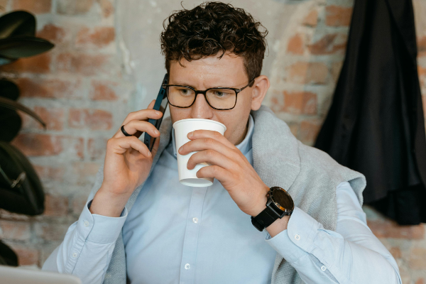 An employee who is often hard to find, sitting at their desk, sipping coffee while looking at their laptop with a phone in hand. Despite being in the office, they appear disengaged and distracted, blending into a laid-back attitude while juggling tasks on their devices.